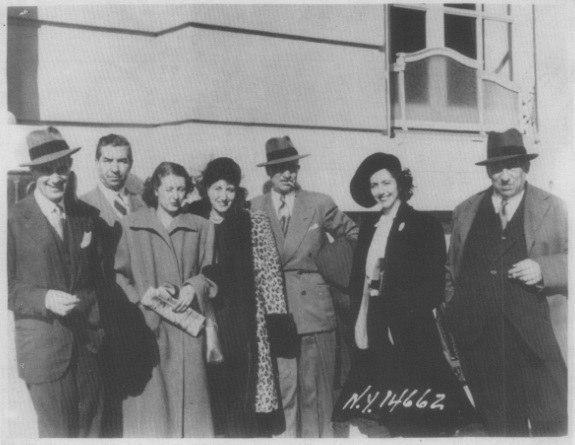 Black and white photograph of seven individuals standing outdoors. They are dressed in formal attire, with hats and coats, posing in a group in front of a building.