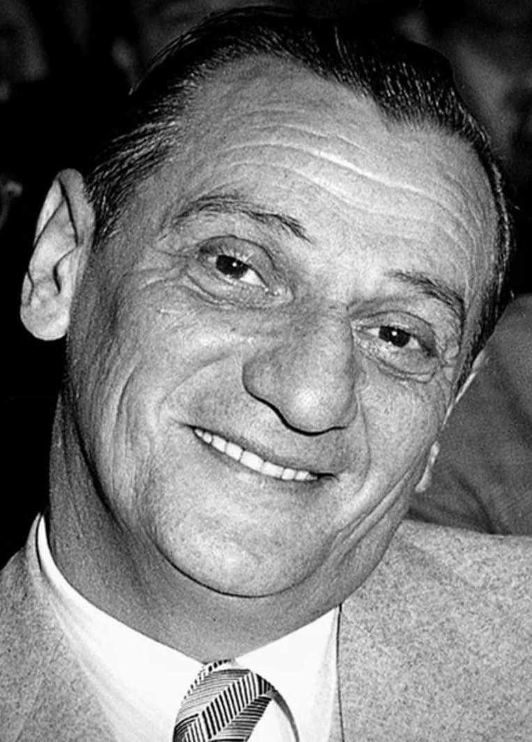A smiling man with slicked-back hair and wearing a light-colored suit and a striped tie.