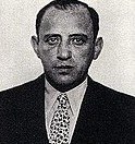 A black and white portrait of a man wearing a dark suit and a patterned tie, looking directly at the camera.