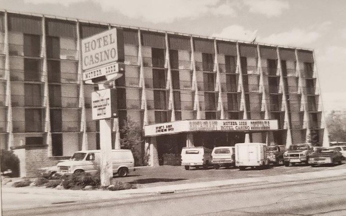 Black and white photo of a mid-century hotel and casino building, featuring a prominent sign that reads 'Hotel Casino.' Several vehicles are parked in front, and the architectural style includes large windows and a covered entrance.
