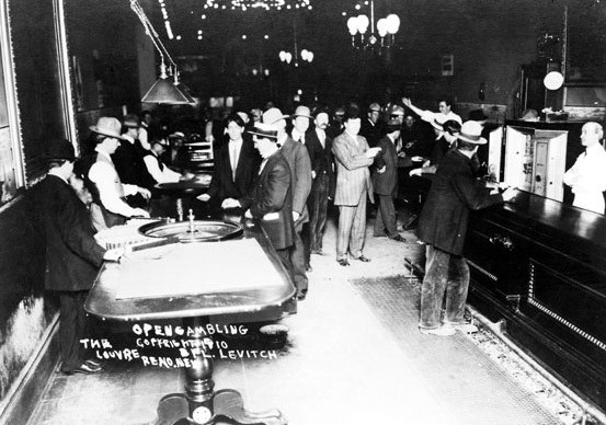 A historic black and white photograph of a bustling gambling hall featuring a roulette table, with people dressed in early 20th-century attire engaging in games. Several patrons are interacting with staff behind a bar, while others stand around the gaming table.