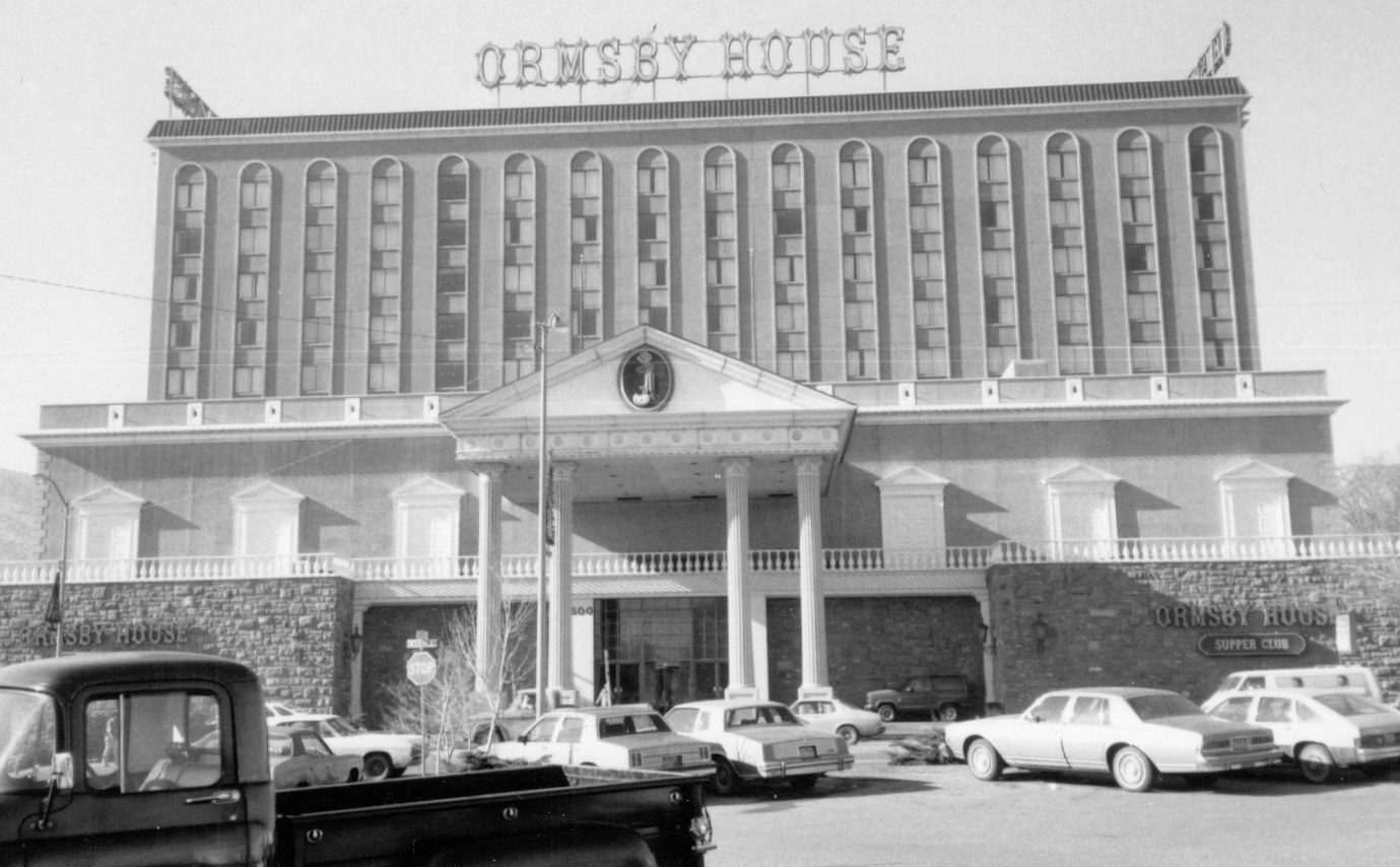 Black and white photo of the Ormsby House hotel featuring a grand entrance with columns and a sign atop the building. Several parked cars are visible in the foreground.