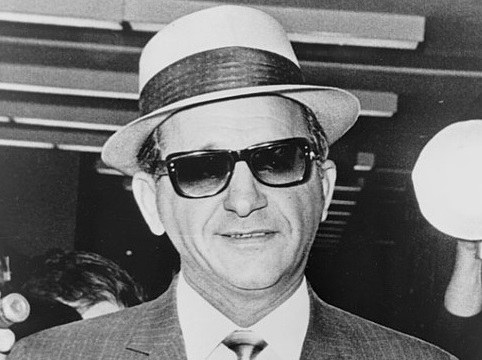 A man in a light-colored suit, white hat, and dark sunglasses, smiling at the camera. He is in an indoor setting with blurred figures in the background.