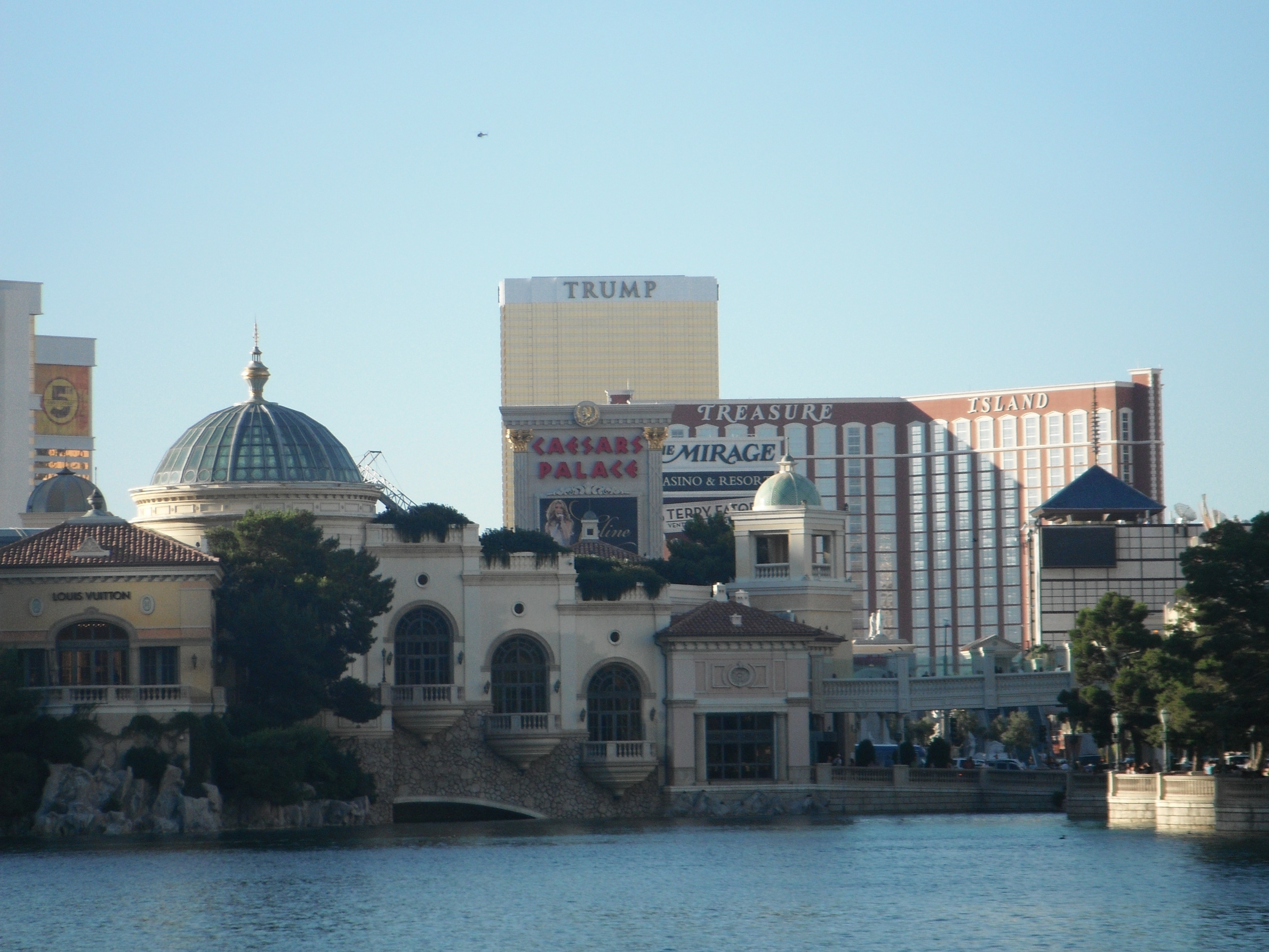 View of Las Vegas Strip featuring Caesars Palace, the Mirage, Treasure Island, and Trump Hotel, with lake and greenery in the foreground.