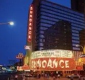 Exterior view of the Sundance theater at night, featuring bright neon lights and a marquee displaying movie titles, with nearby fast food restaurants visible.