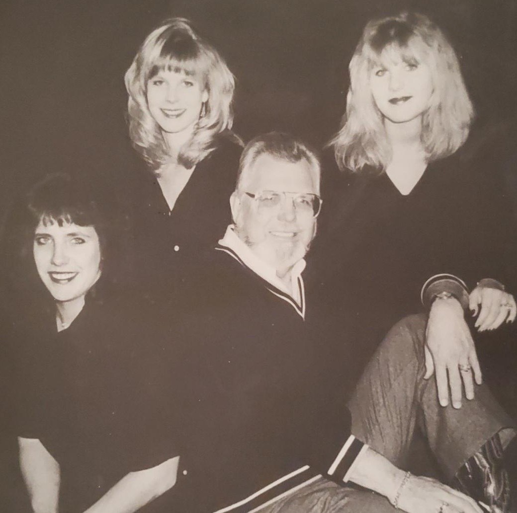 A black and white photo of four individuals posing together. There are three women with varying hairstyles and one man sitting in the front. They are all wearing dark clothing and smiling at the camera.