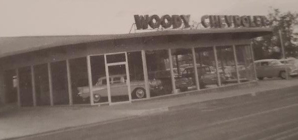 A vintage Chevrolet car dealership called 'Woody Chevrolet', featuring large glass windows displaying classic cars for sale.