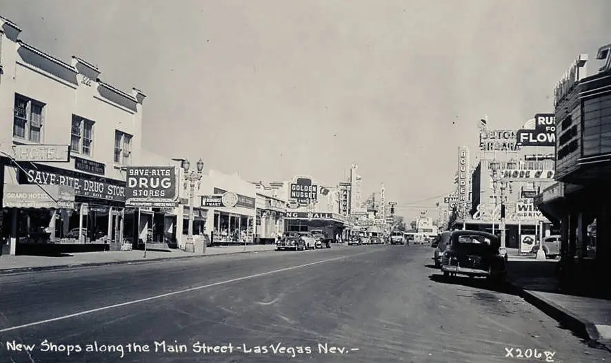 Historic view of downtown Las Vegas, showing storefronts, casino signs and cars along the street.