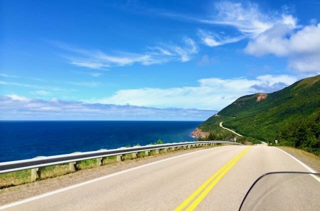 A winding coastal road between a green hillside and the blue ocean