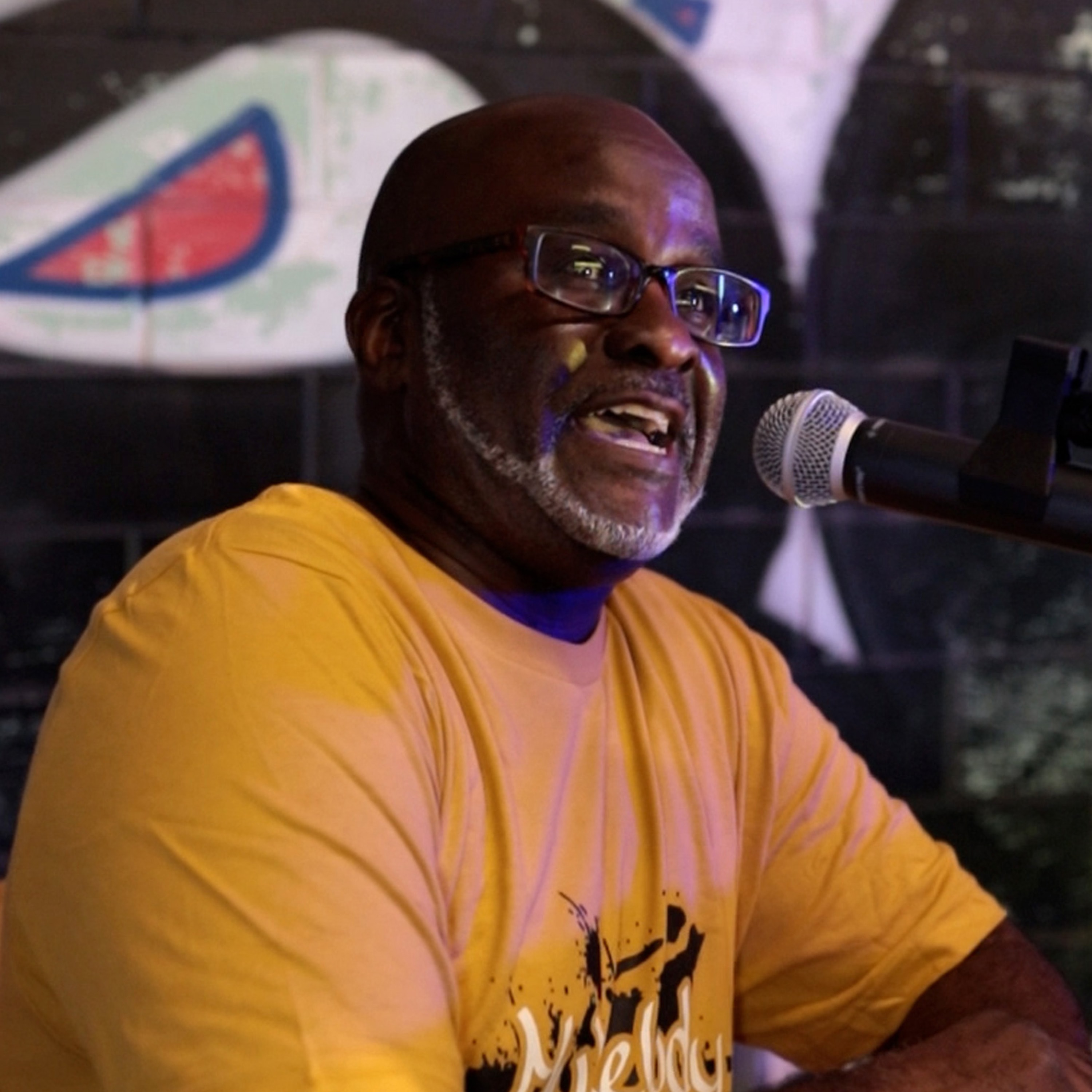 Tyrine the Artist speaking into a microphone while wearing the My Melody Is Motivational gold shirt