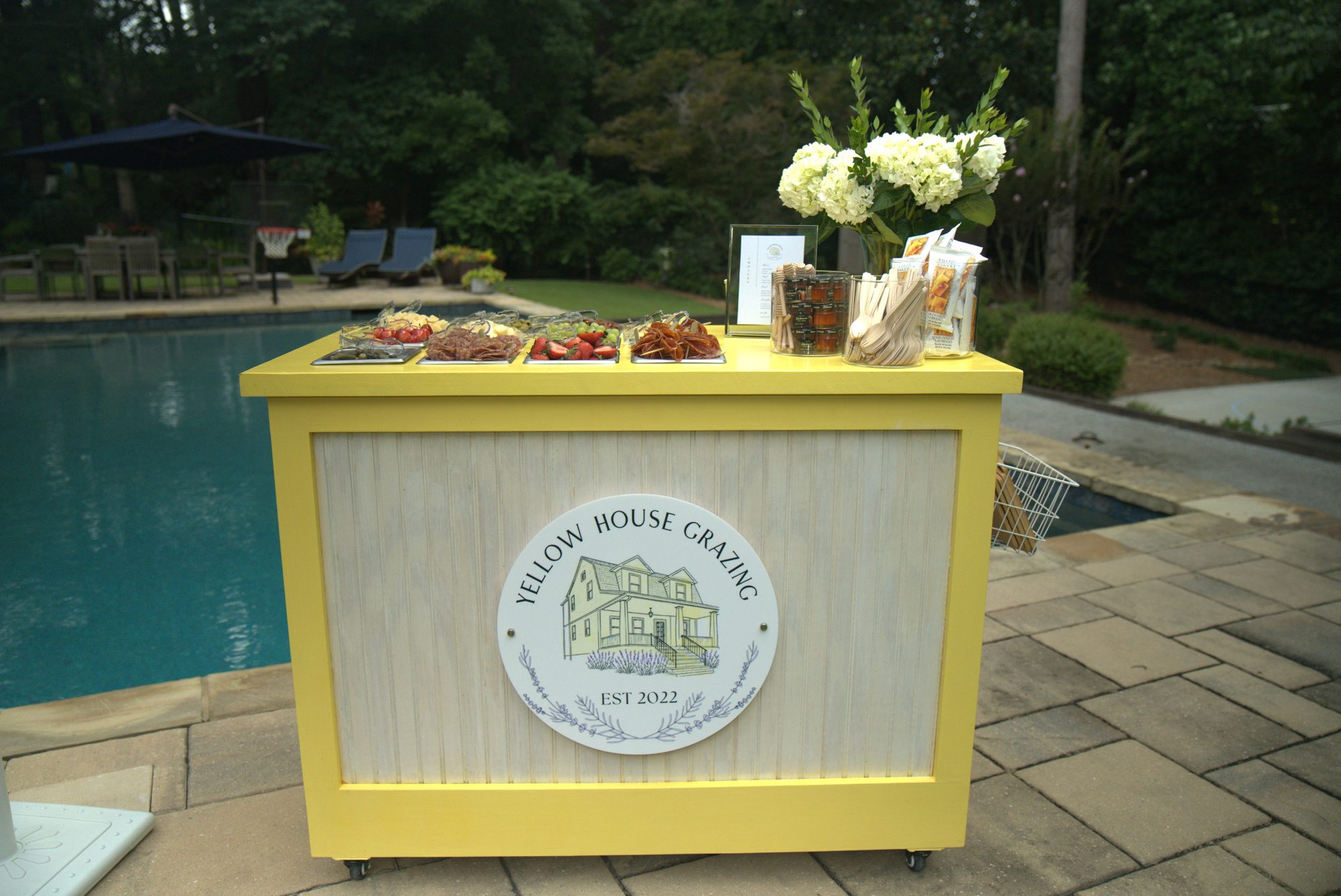 Yellow House Grazing’s yellow cart set beside a pool with flowers and a grazing spread