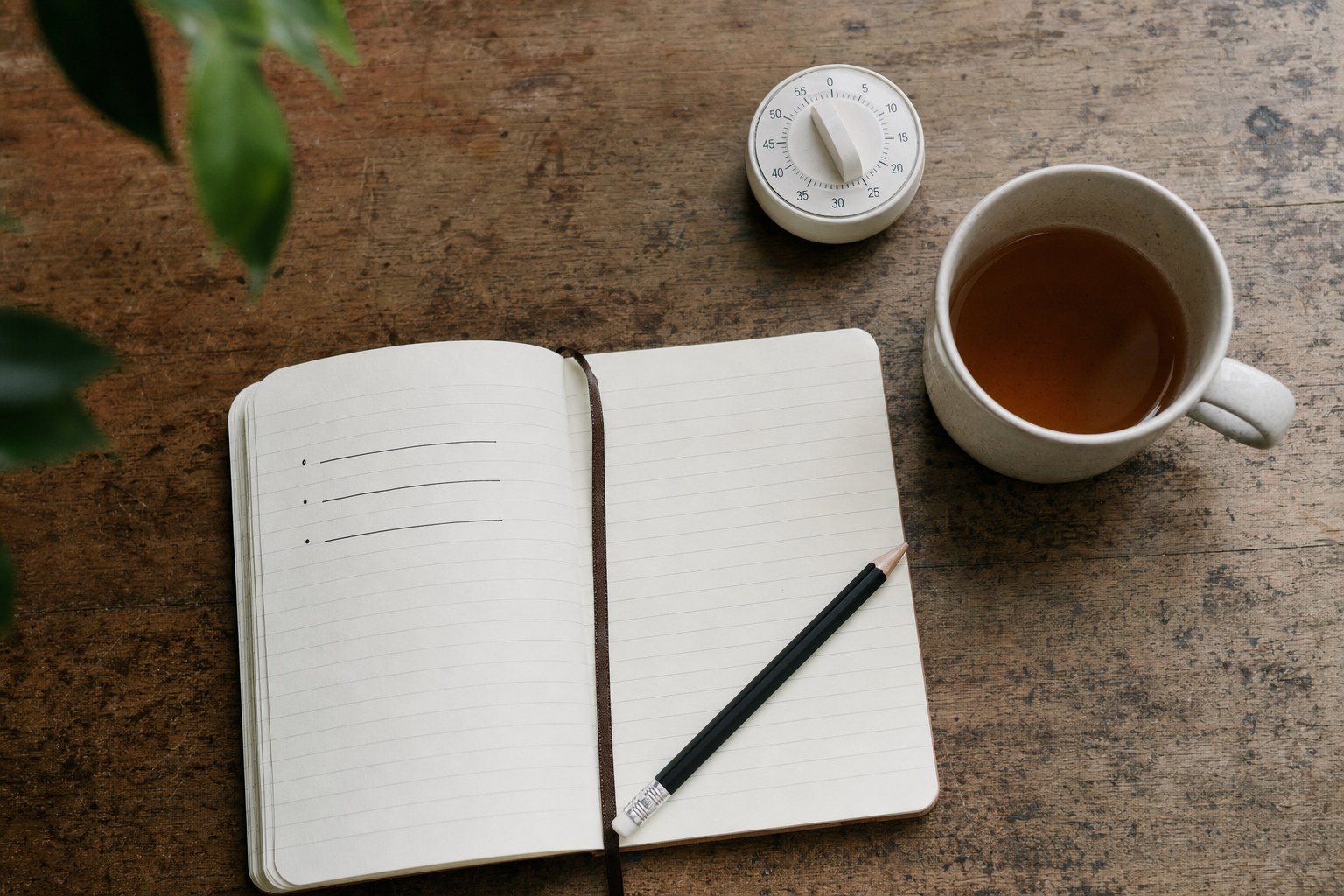 An open notebook, kitchen timer, pencil, and cup of tea on a wooden table