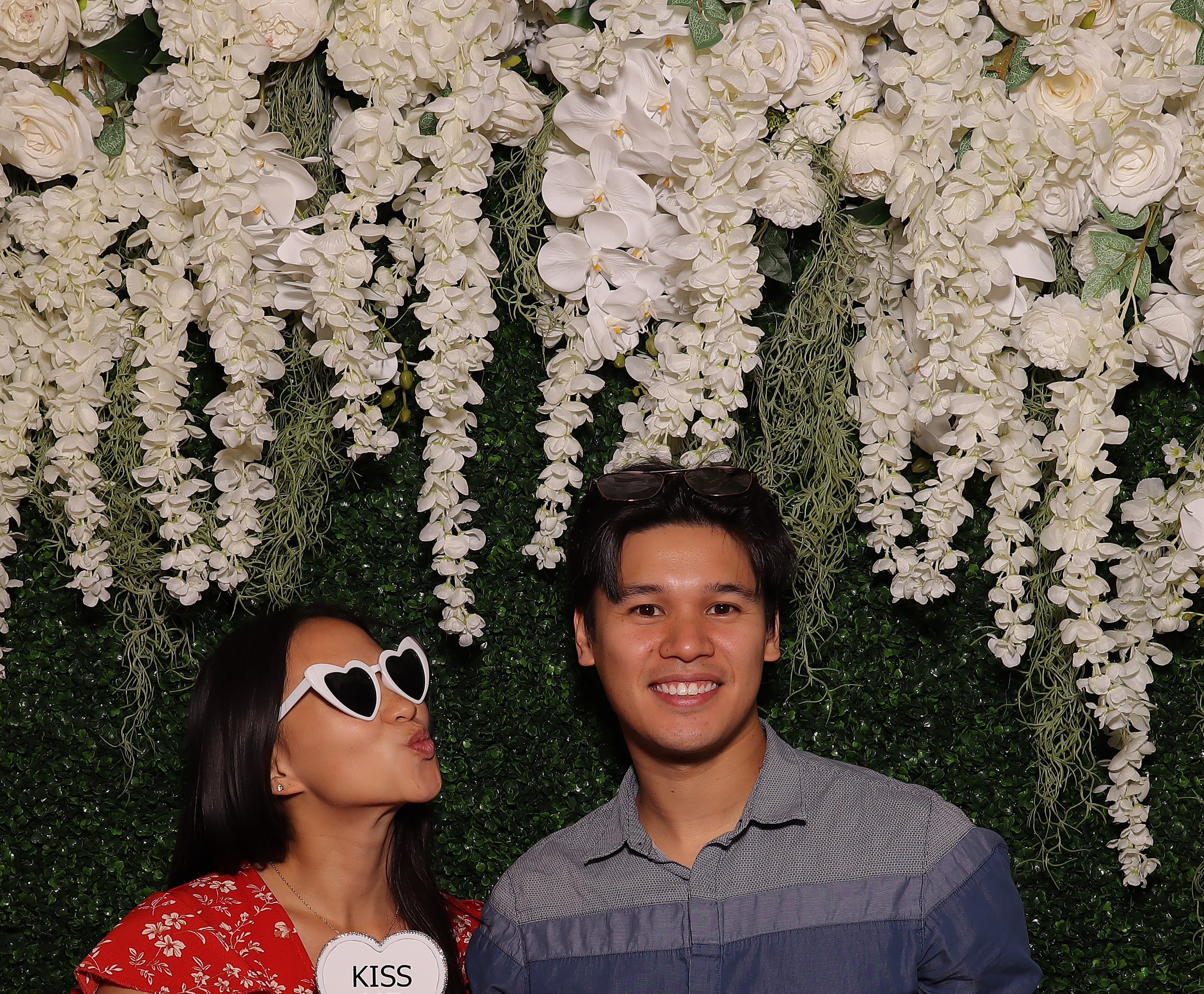 Two guests having fun in front of a white floral and greenery Photo Booth backdrop