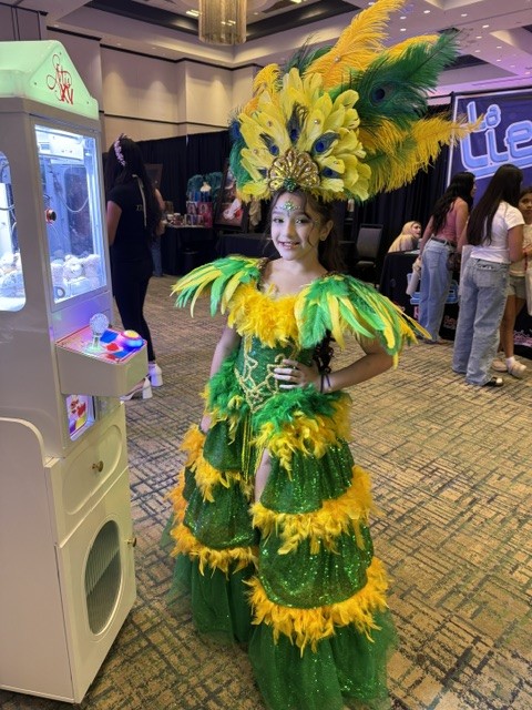 Performer in green and yellow costume beside The Pose Garden claw machine at a Phoenix-area quinceañera expo