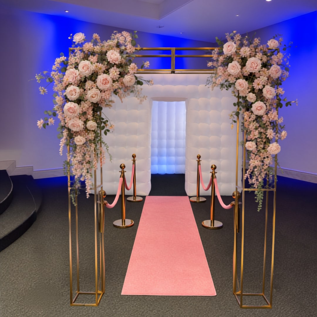 White inflatable photo booth enclosure with a gold floral arch and pink carpet
