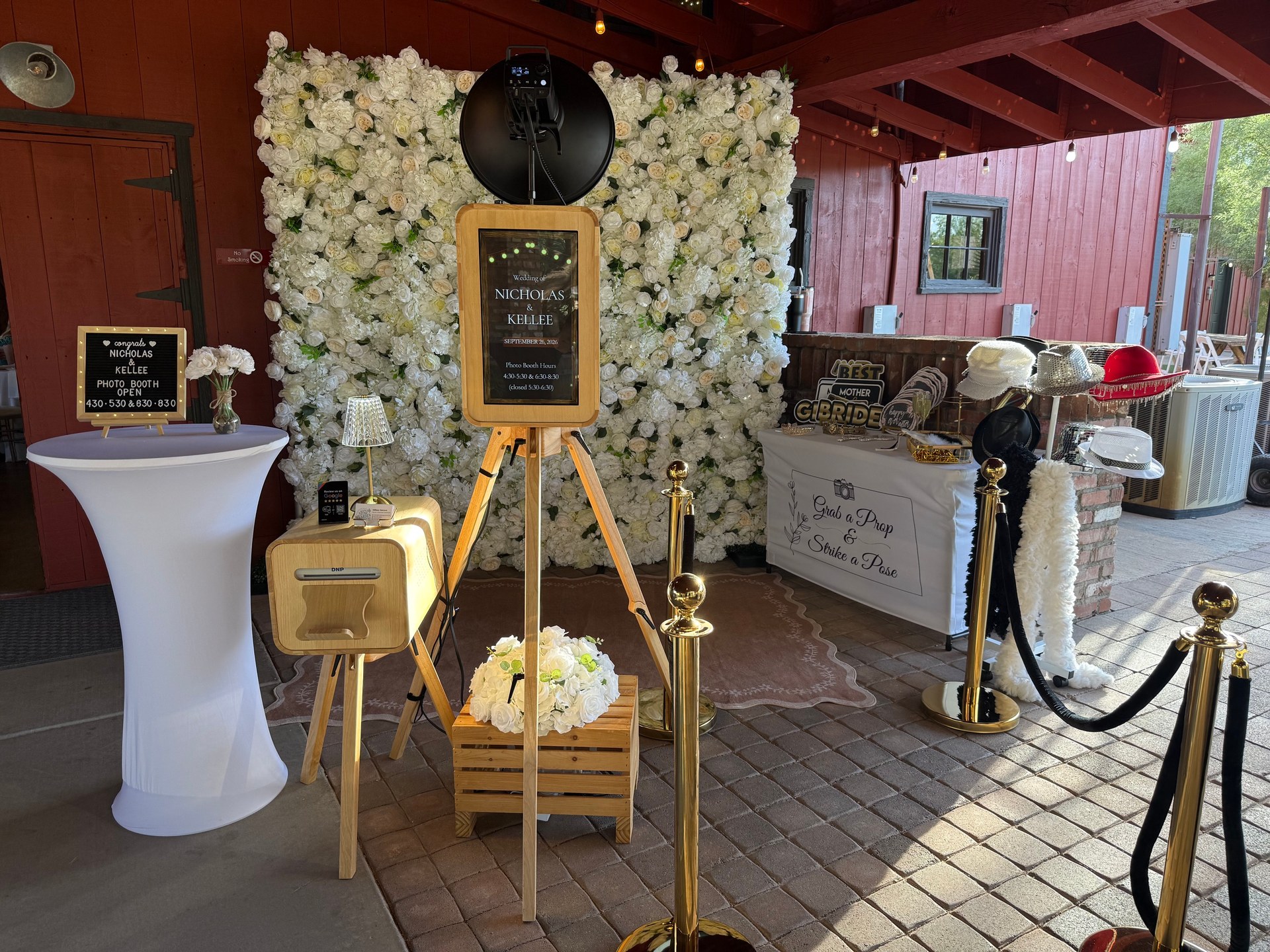 The Artisan oak Photo Booth with a white flower wall, printer and props at a celebration