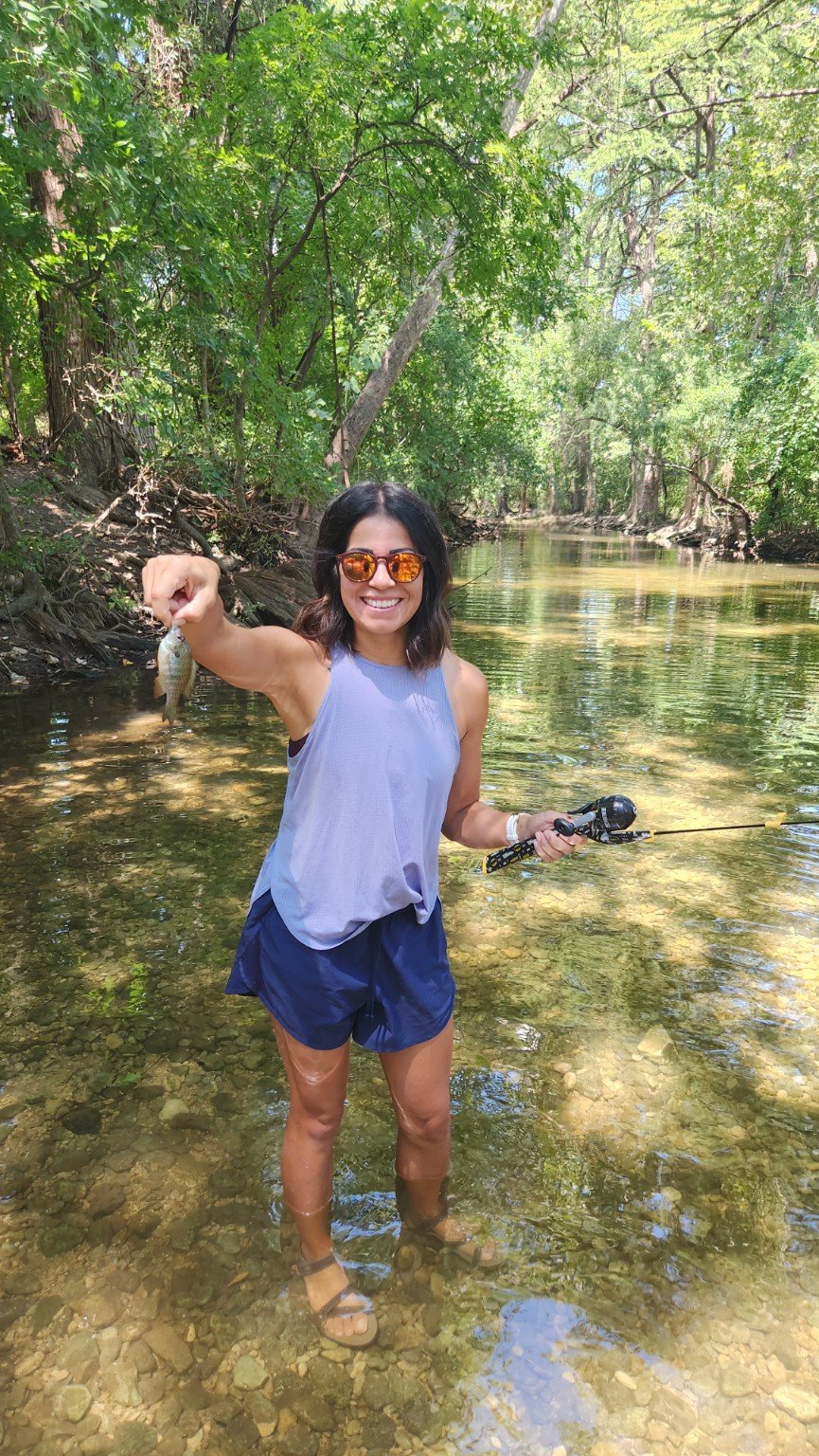 Roxanne wearing a lavender top and holding a small fish in a tree-lined creek