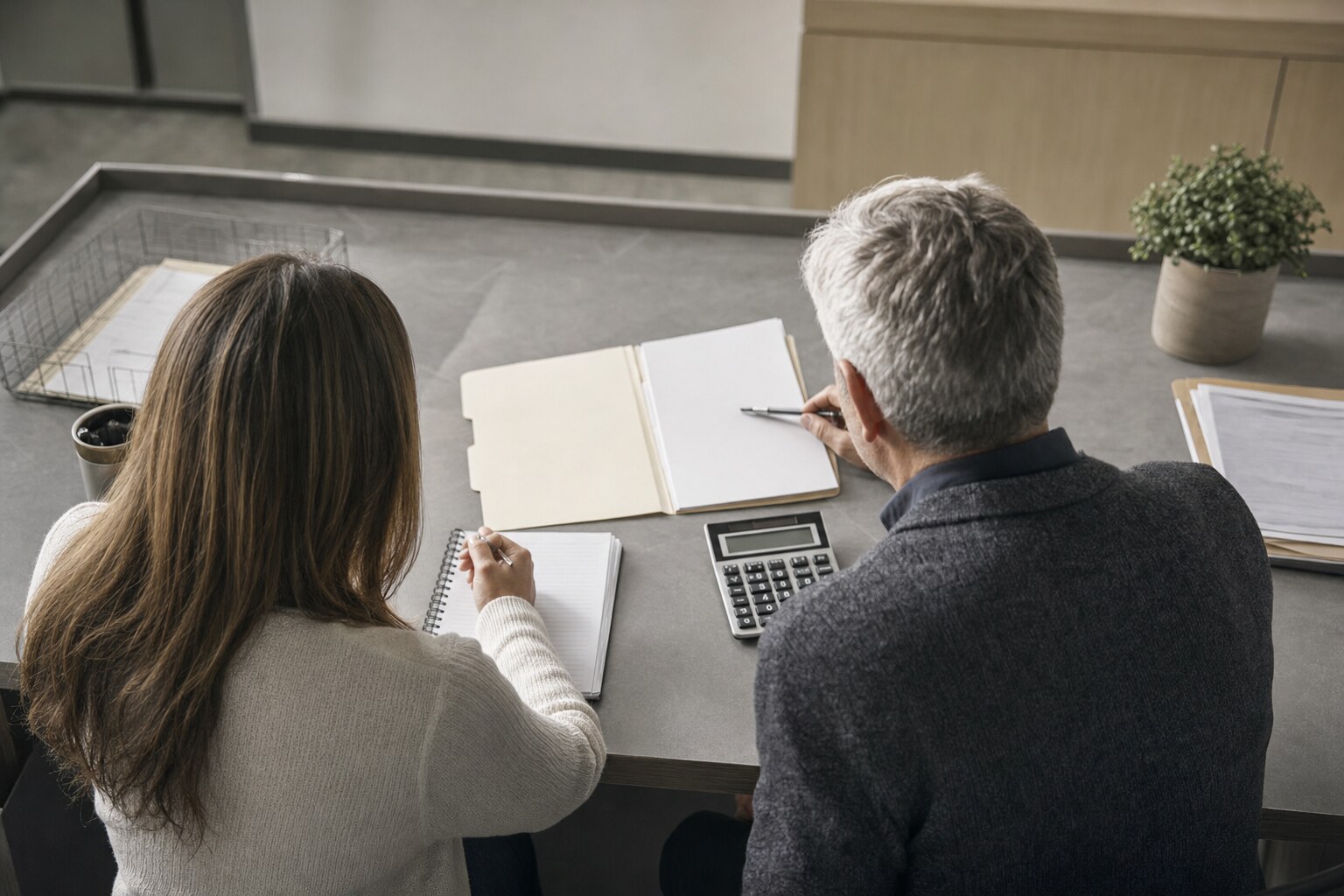 Two people reviewing a plain folder, notebook, and calculator across an office desk