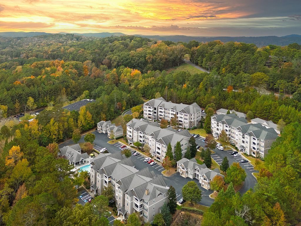 Aerial view of apartment complex nestled in lush, colorful autumnal trees; parking lot, pool visible.