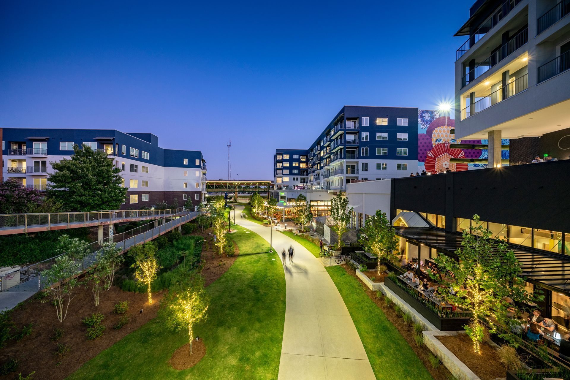 Night view of a pedestrian walkway lined with buildings and greenery. People stroll.