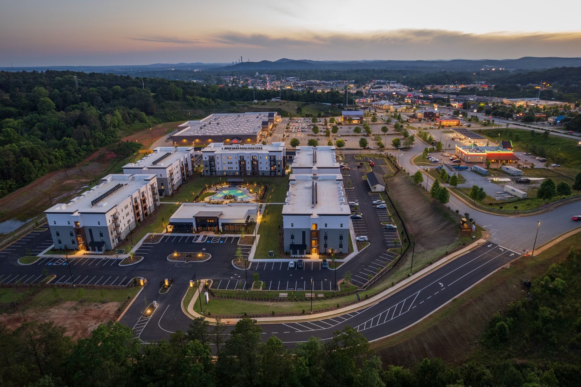 Aerial view of modern apartment complex, pool, and shopping center at dusk with a city in the background.