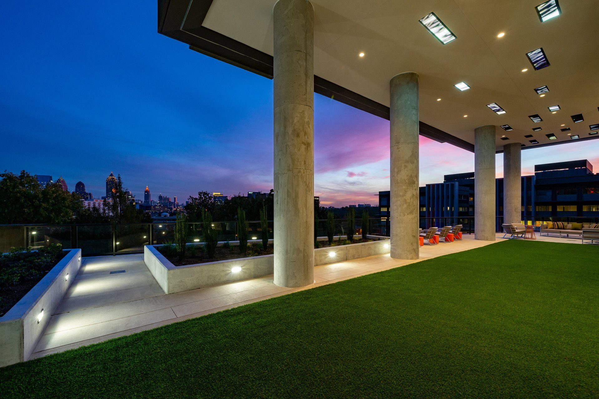 Rooftop patio with green turf, concrete columns, and a city skyline at dusk with pink and blue sky.