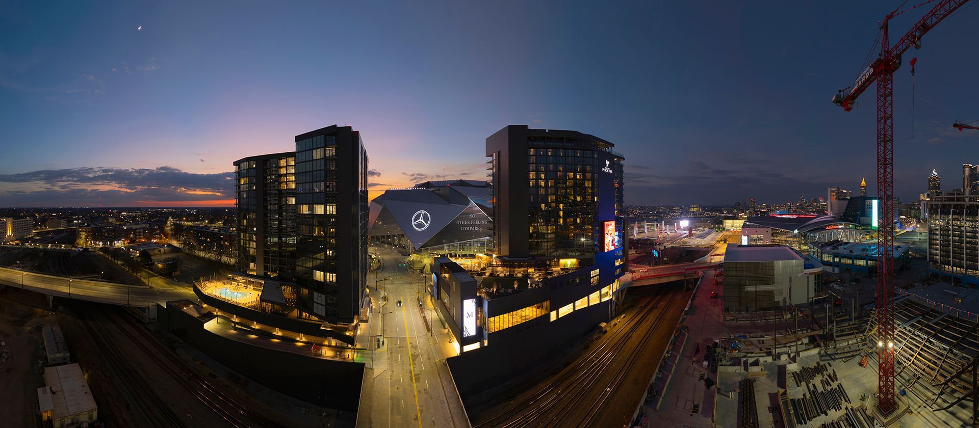 Panoramic cityscape at dusk, featuring lit buildings, a sports stadium, and construction cranes.