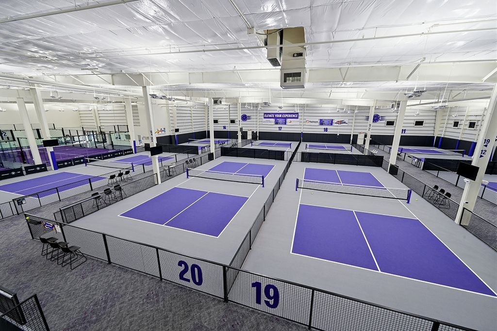 Indoor pickleball courts, gray floors, purple playing areas, metal fences, overhead lighting.