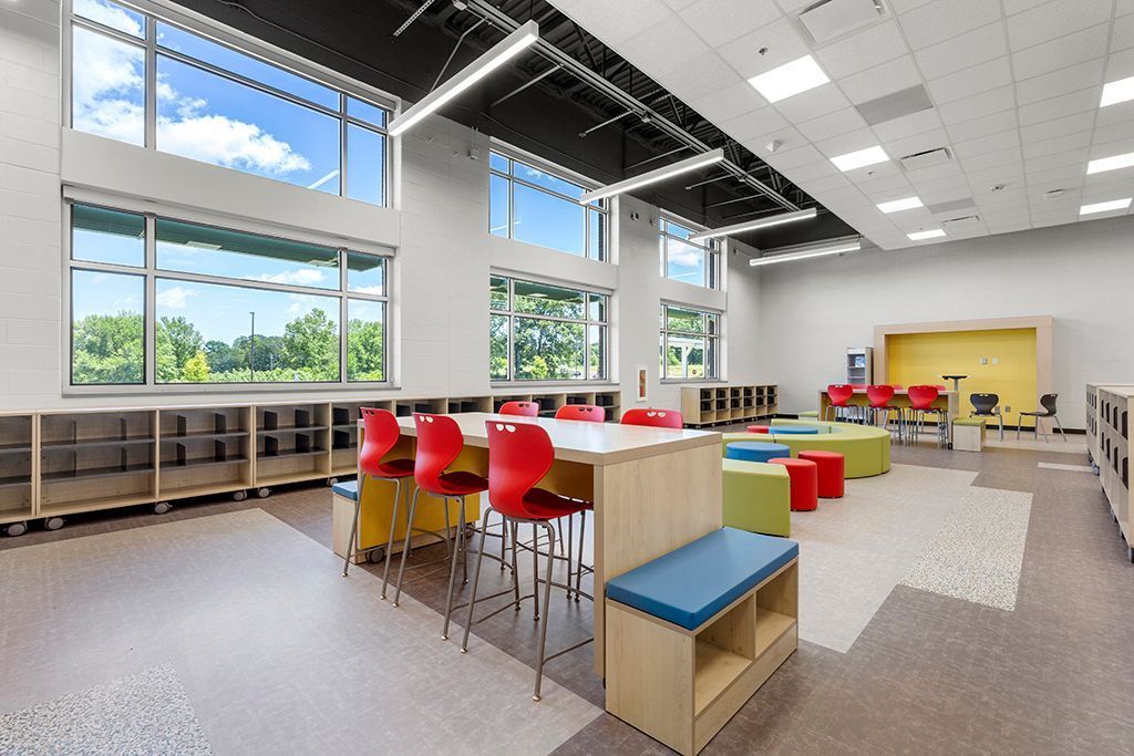 Bright classroom with tables, chairs, and colorful seating areas near large windows overlooking a green landscape.
