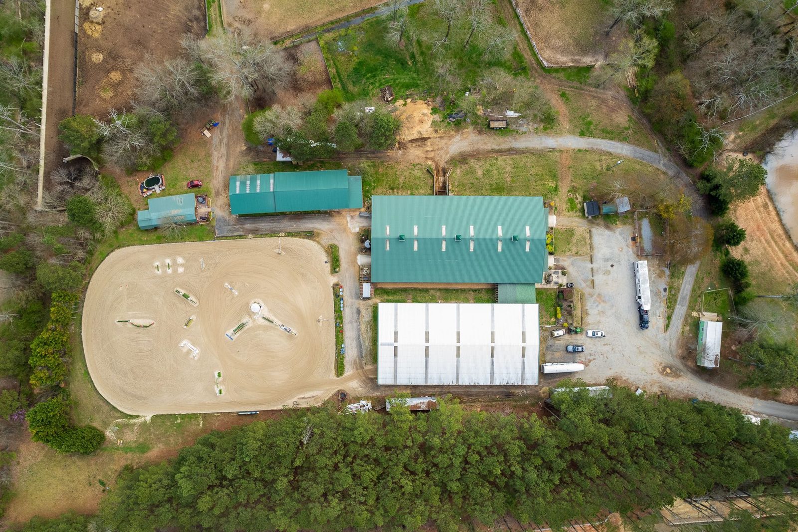Aerial view of a horse farm with an arena, buildings with green roofs, and surrounding trees.