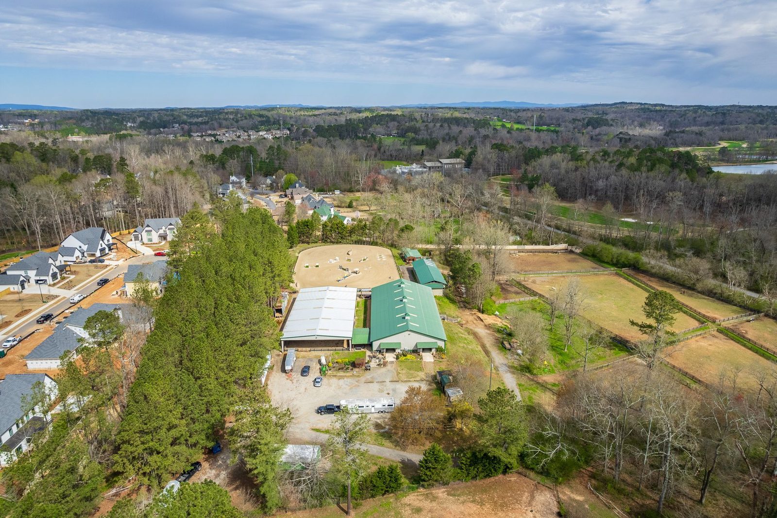 Aerial view of a horse stable with covered riding arenas, fenced pastures, and houses in the distance.