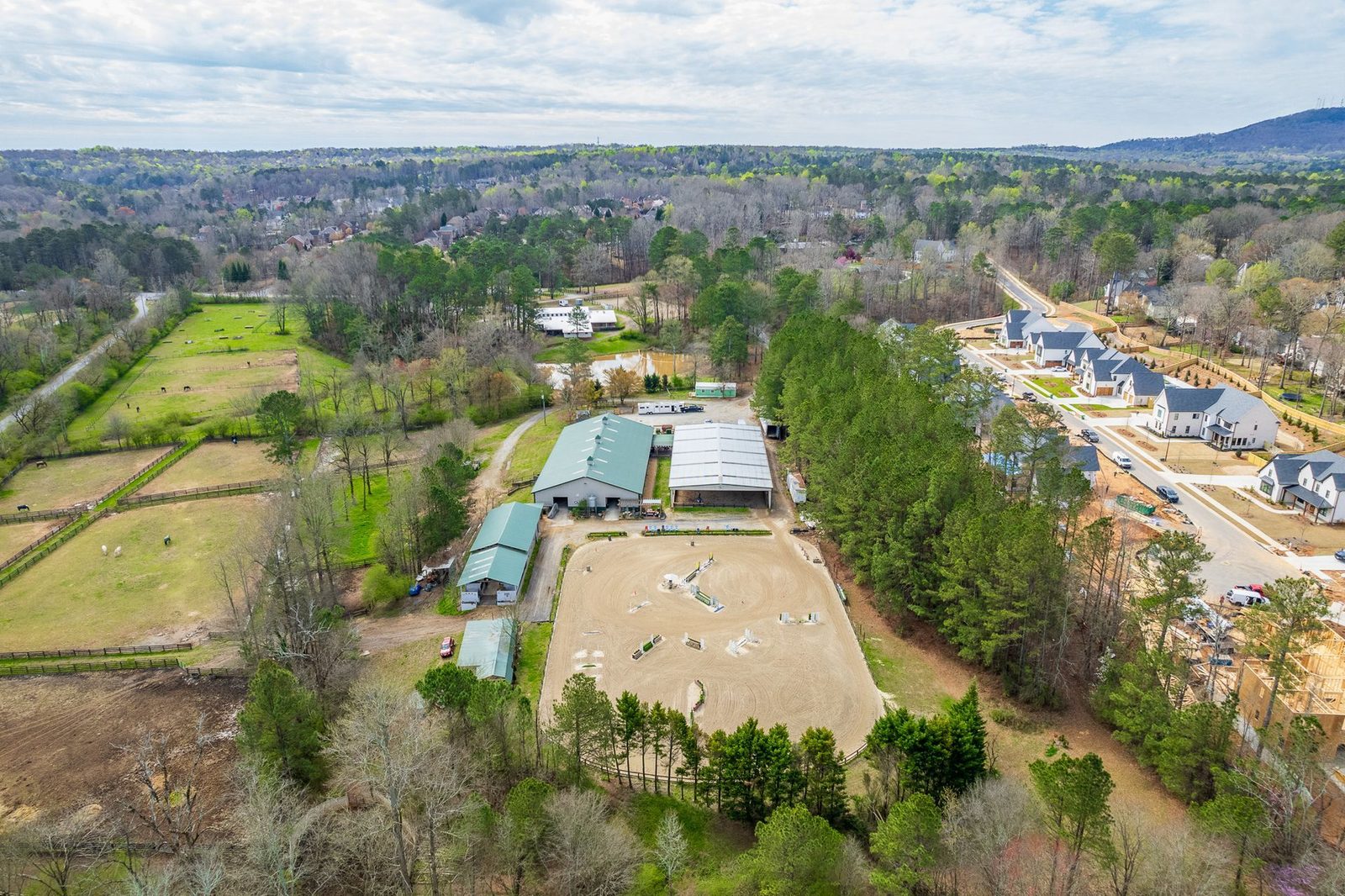 Aerial view of an equestrian facility with riding arena, stables, and nearby houses. Green trees surround the area.