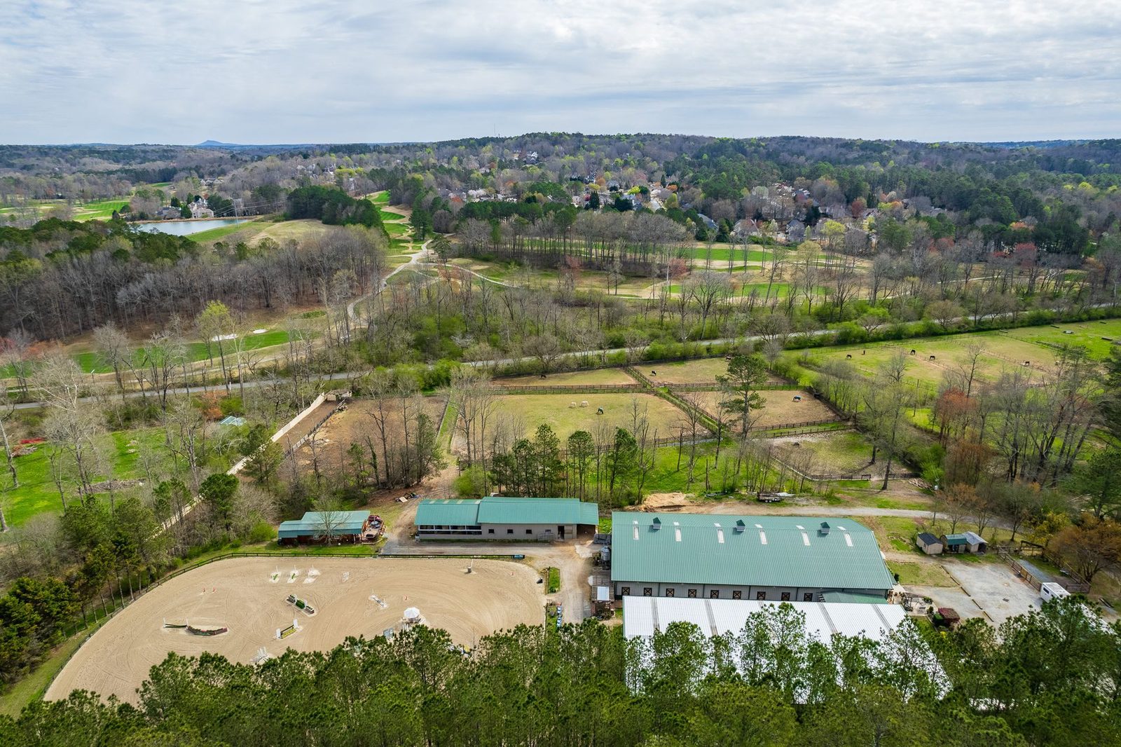 Aerial view of an equestrian facility with riding arenas, stables, and surrounding wooded landscape.