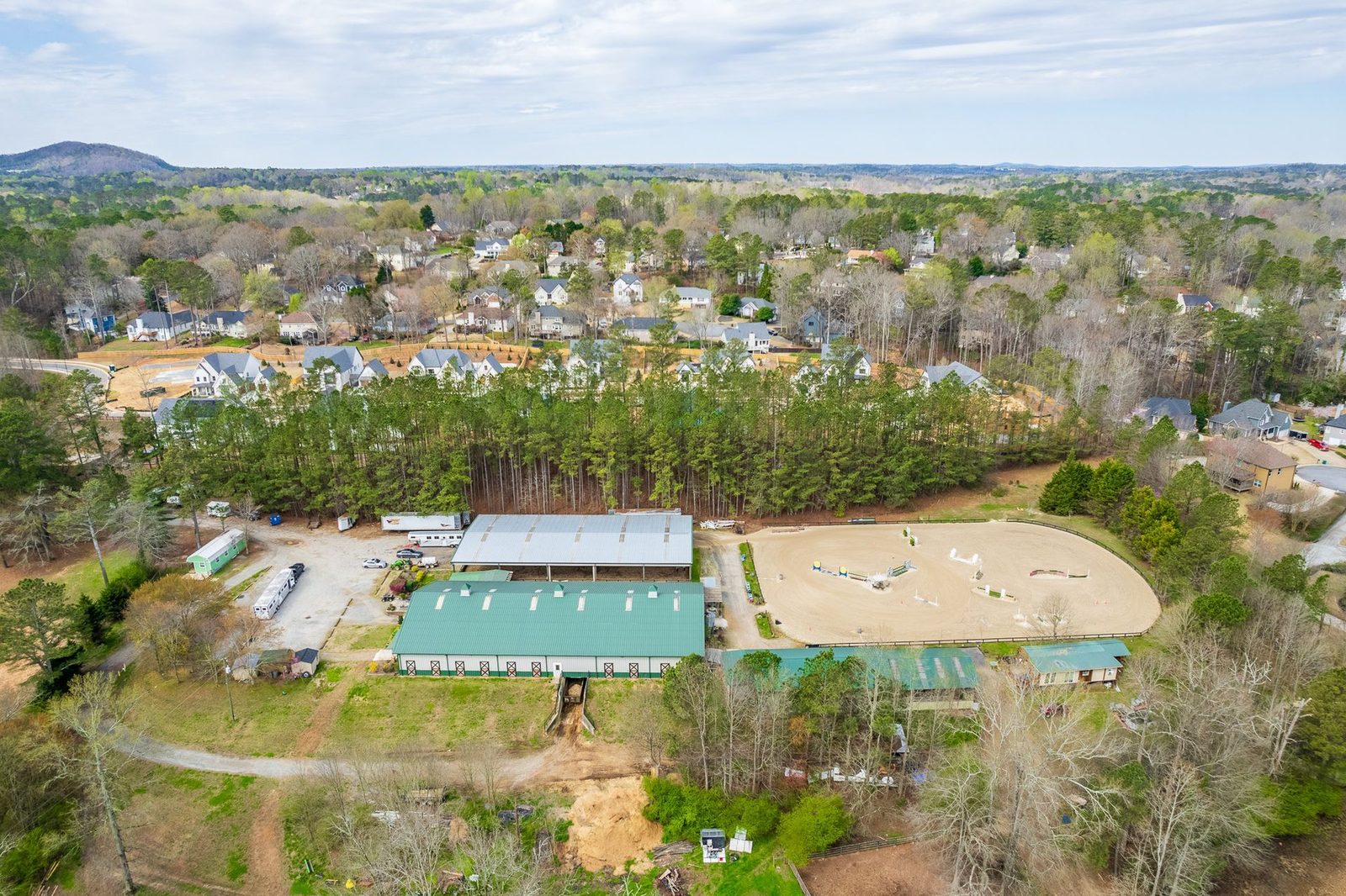 Aerial view of a horse stable and riding arena. Green roof, light-colored arena with jumps, surrounded by trees and houses.