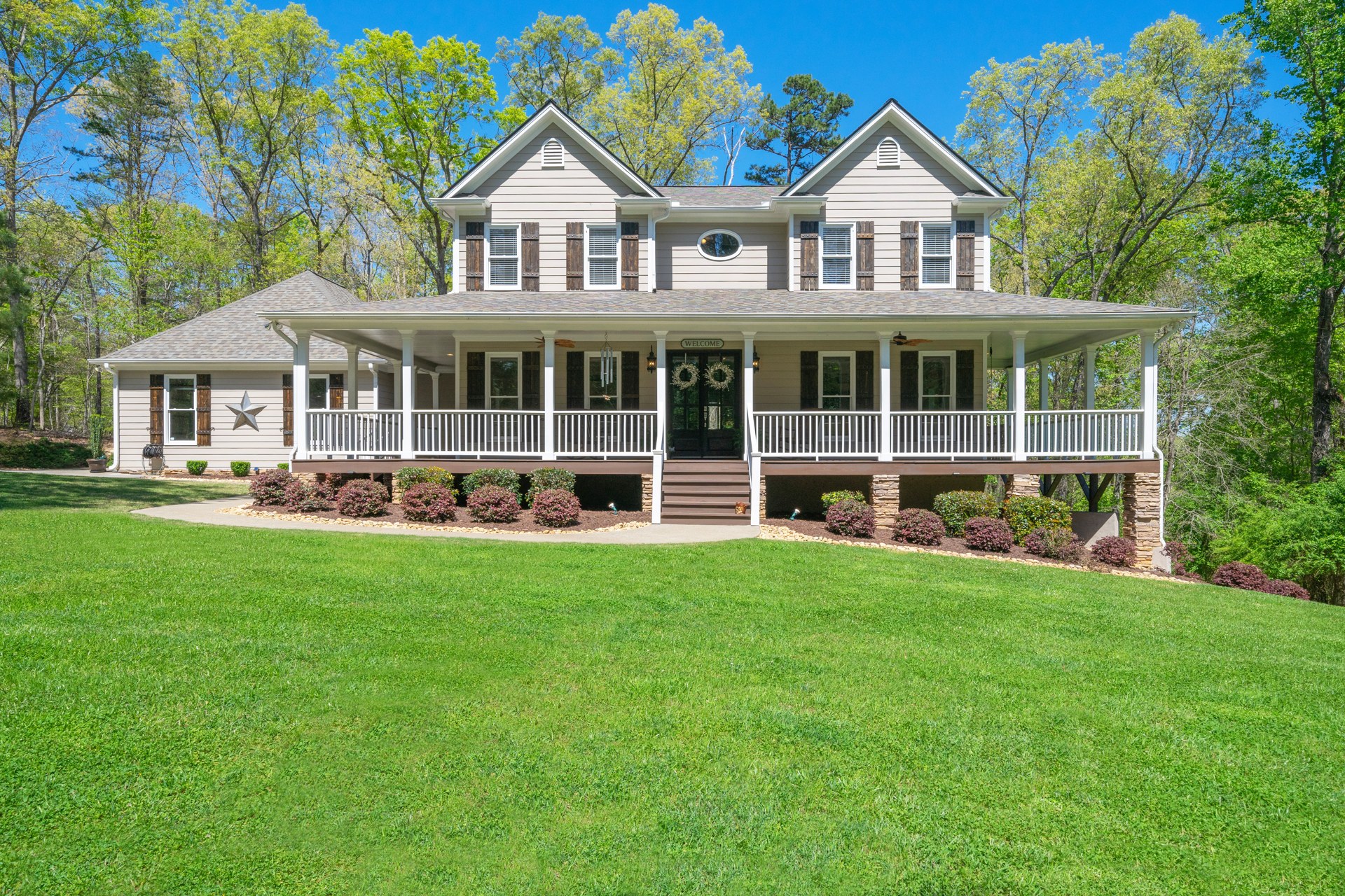 Two-story Ball Ground home with a broad wraparound porch, wood shutters, and a green lawn surrounded by trees