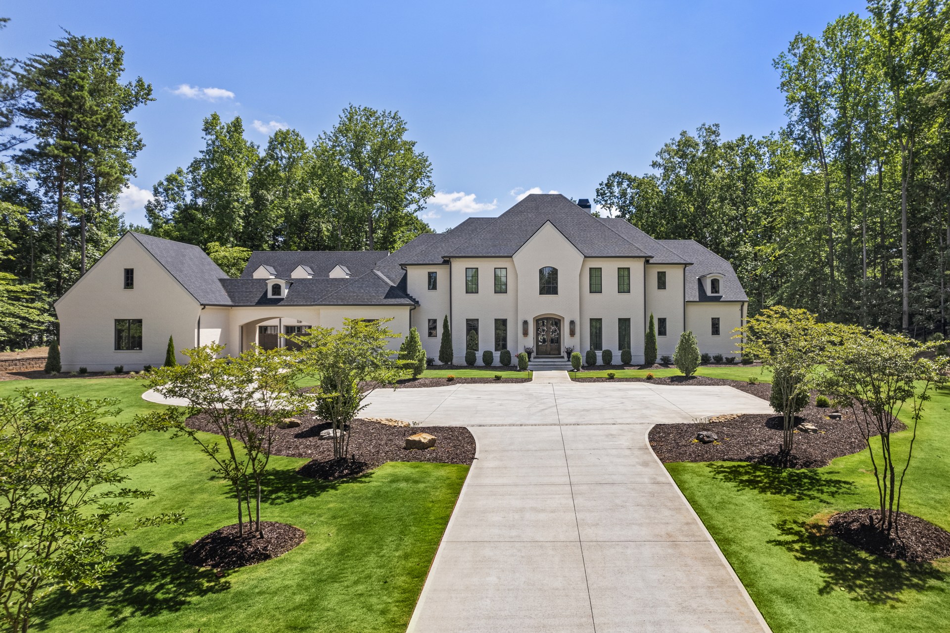 Large white home with a dark roof, circular driveway, manicured lawn, and wooded surroundings