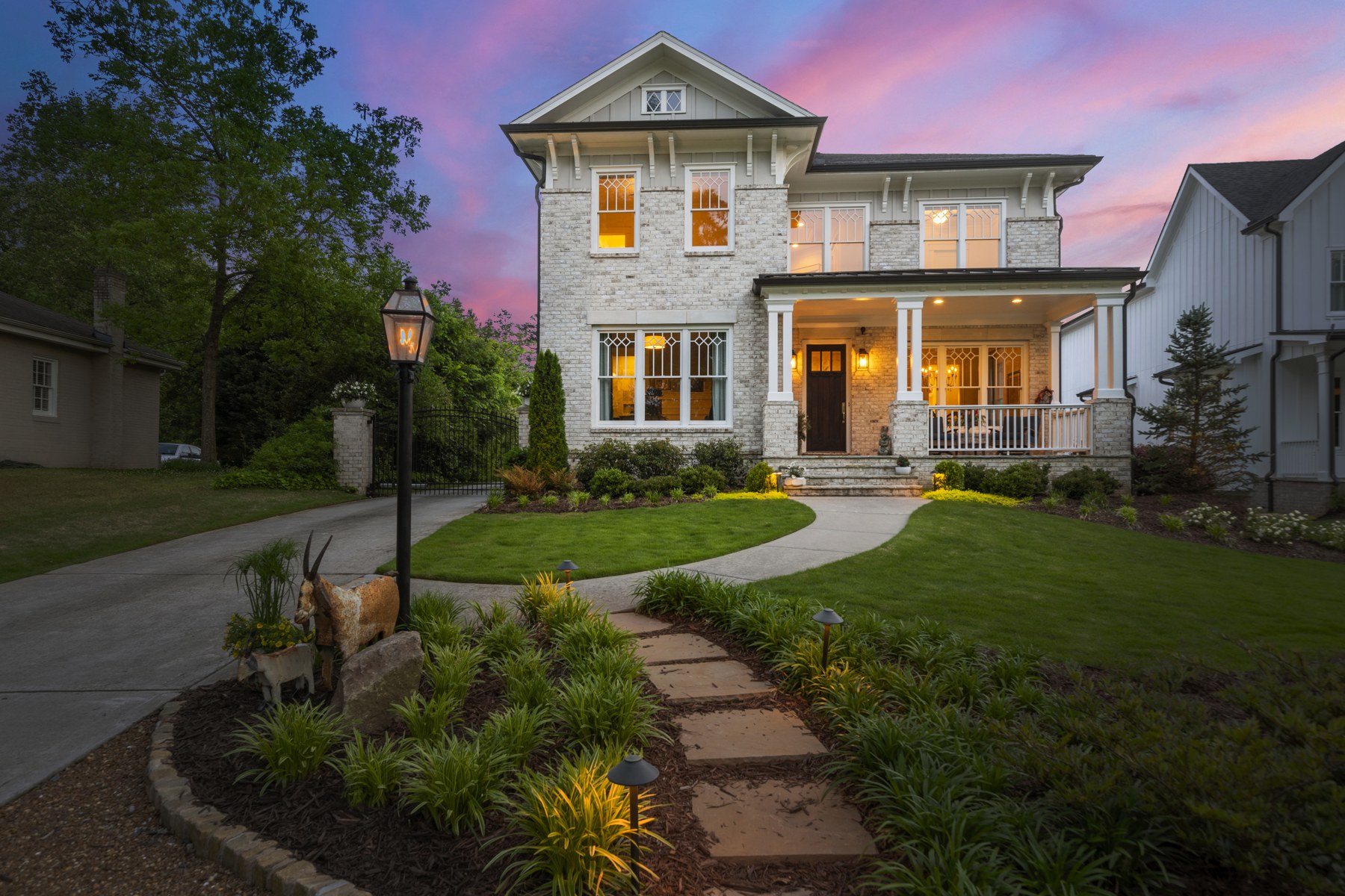 White brick home at twilight with a lit front porch, landscaped garden, and pink evening sky