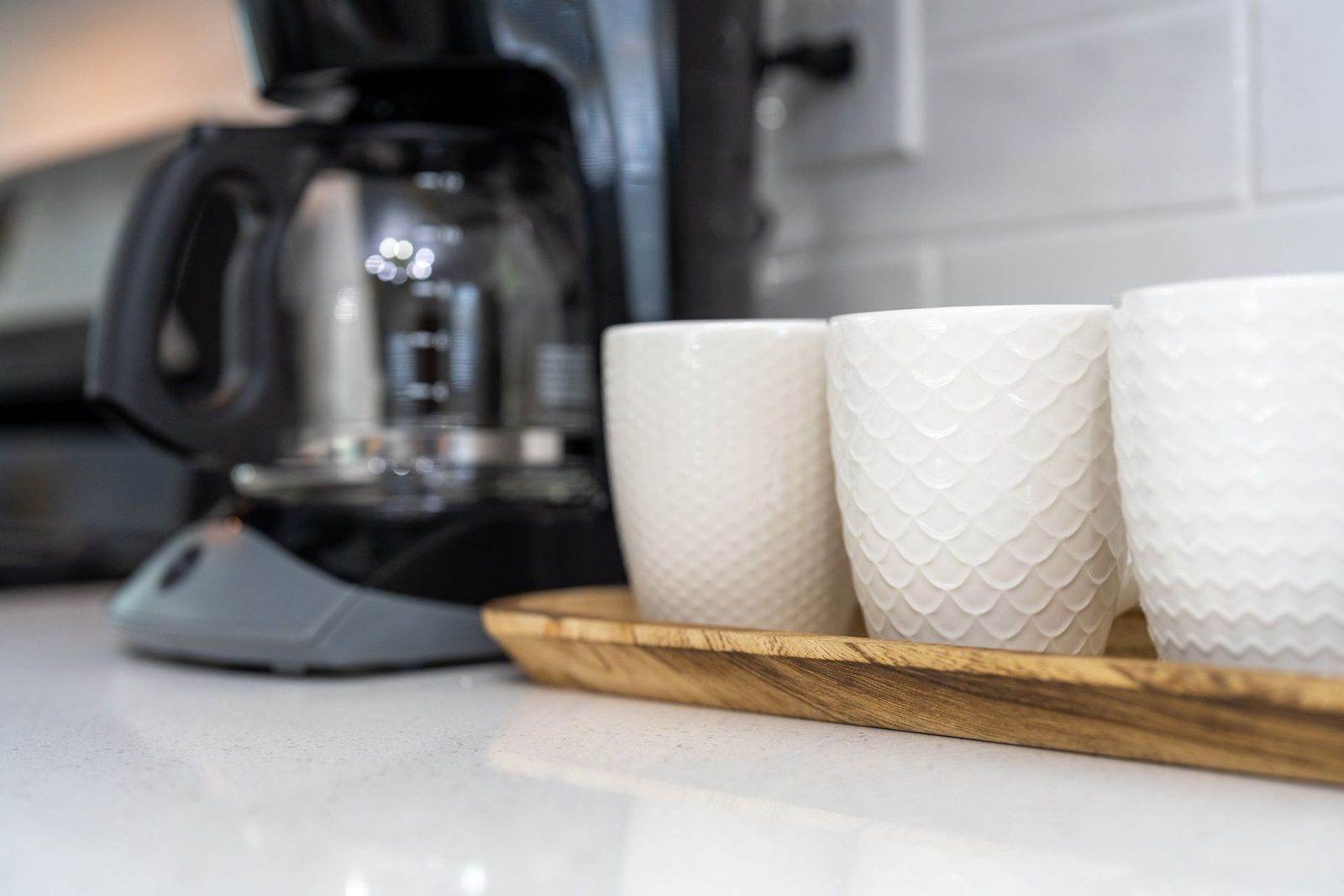 Three white cups are sitting on a wooden tray on a counter next to a coffee maker.