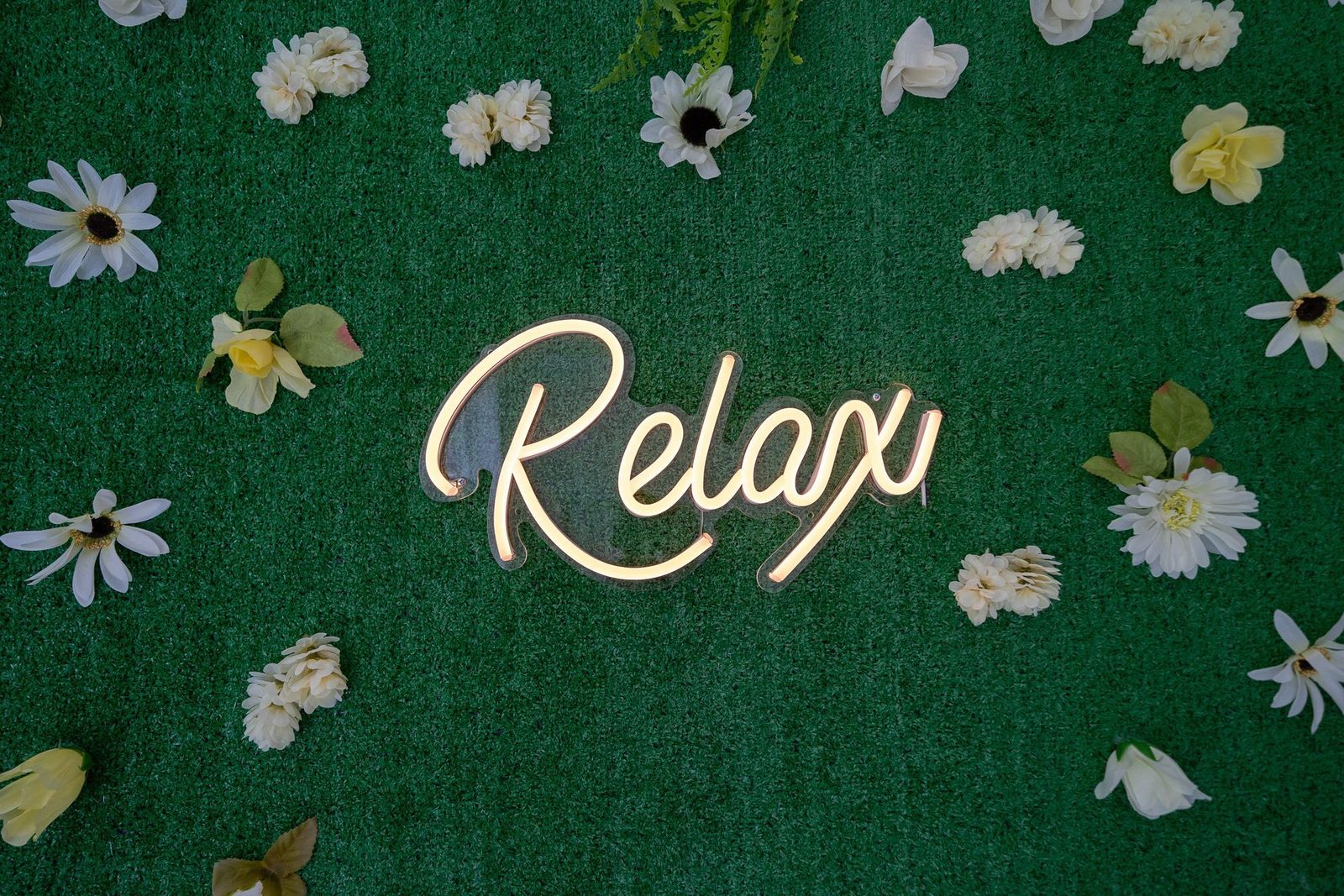 A neon sign that says `` relax '' is surrounded by flowers on a green field.