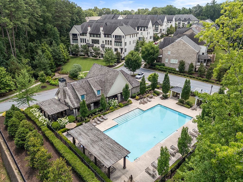 Aerial view of a community pool and clubhouse surrounded by buildings and trees on a sunny day.