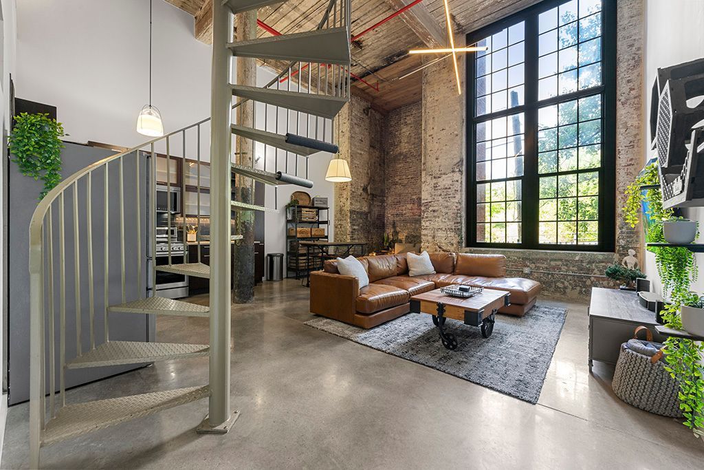 Loft interior with a spiral staircase, leather sectional, and large window. Exposed brick and concrete floors.