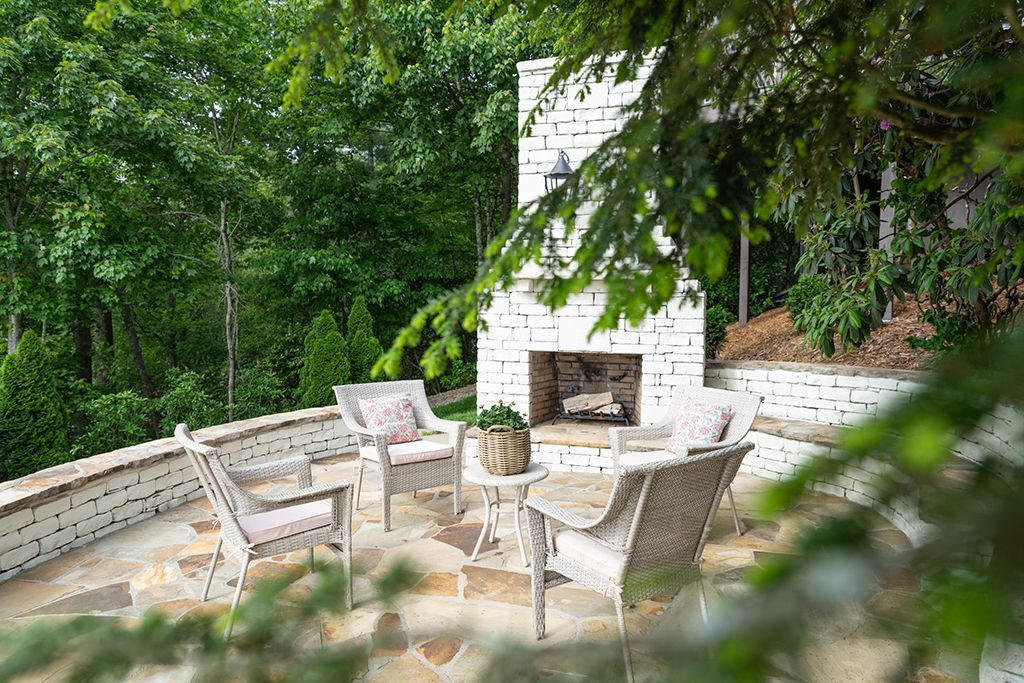 Patio with fireplace, seating, and lush greenery. White brick, stone, and woven furniture create a cozy outdoor space.