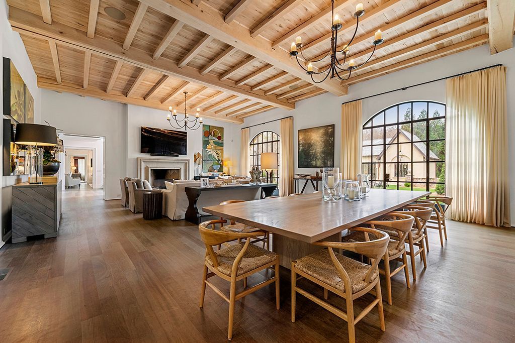 Spacious dining room with long table, wooden floor, and arched windows. Exposed wood ceiling, chandelier, and modern decor.