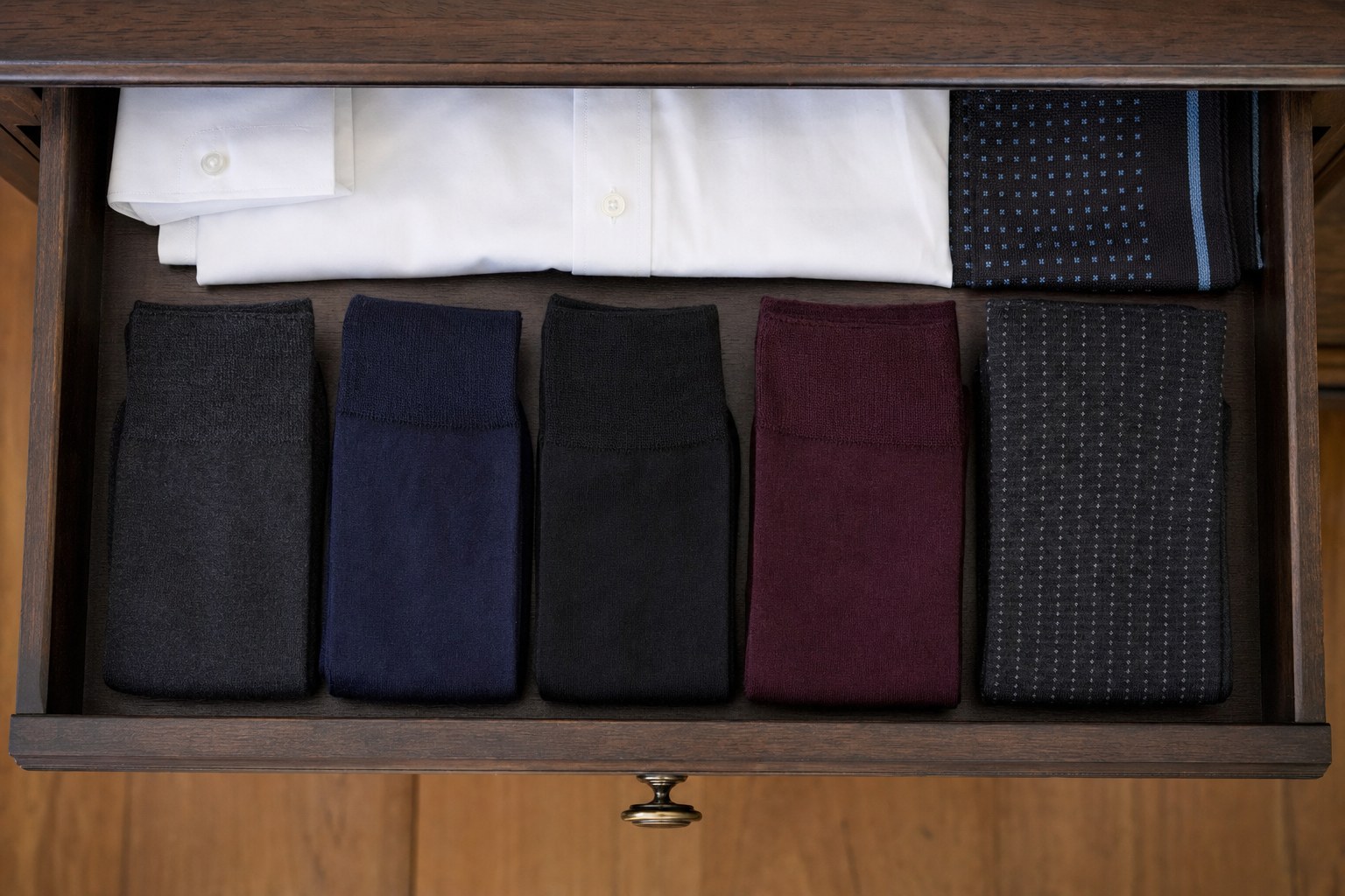 A wood dresser drawer with charcoal, navy, black, burgundy, and subtly patterned dress socks arranged beside a folded white shirt