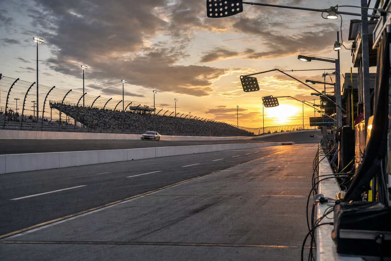 An unbranded stock car passing an oval-race pit lane at sunset