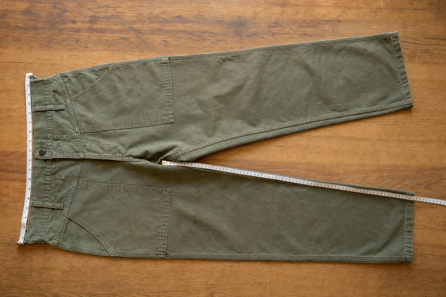Olive canvas work pants laid flat with a measuring tape at the waist and inseam
