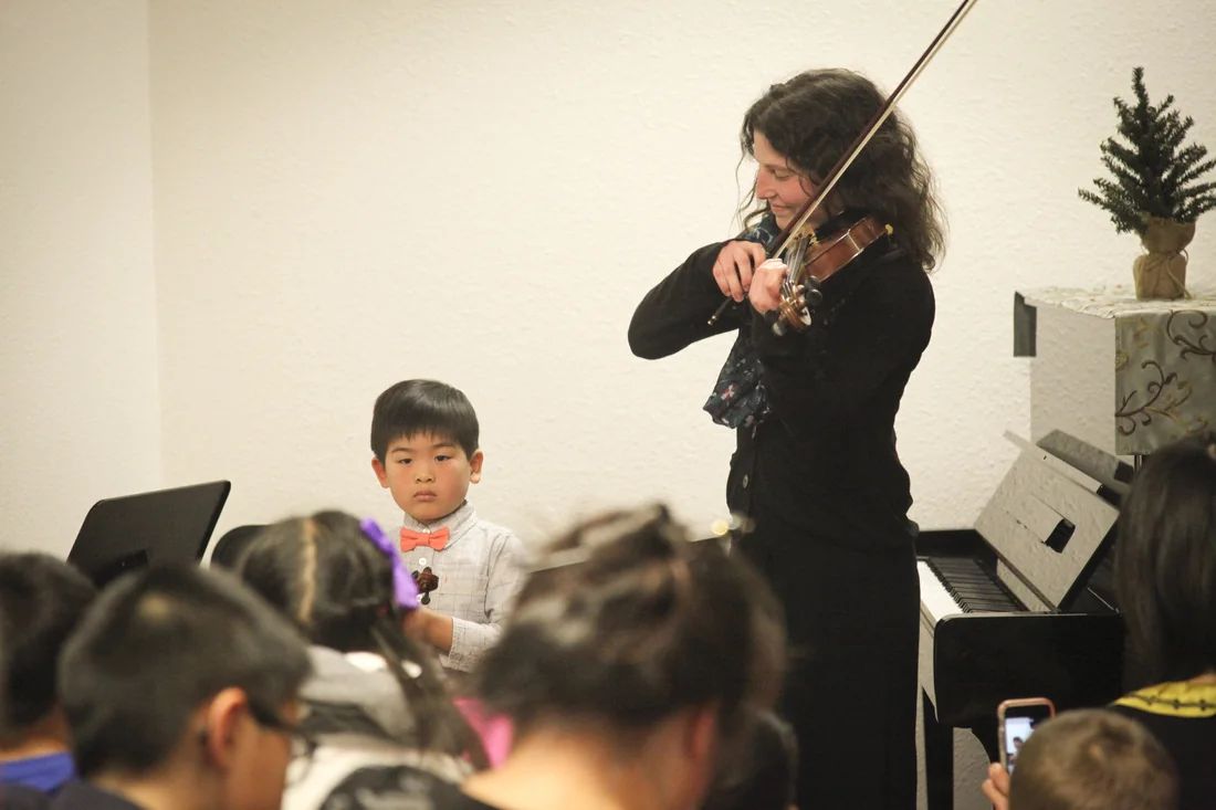 Violin teacher performing beside a young student in front of families