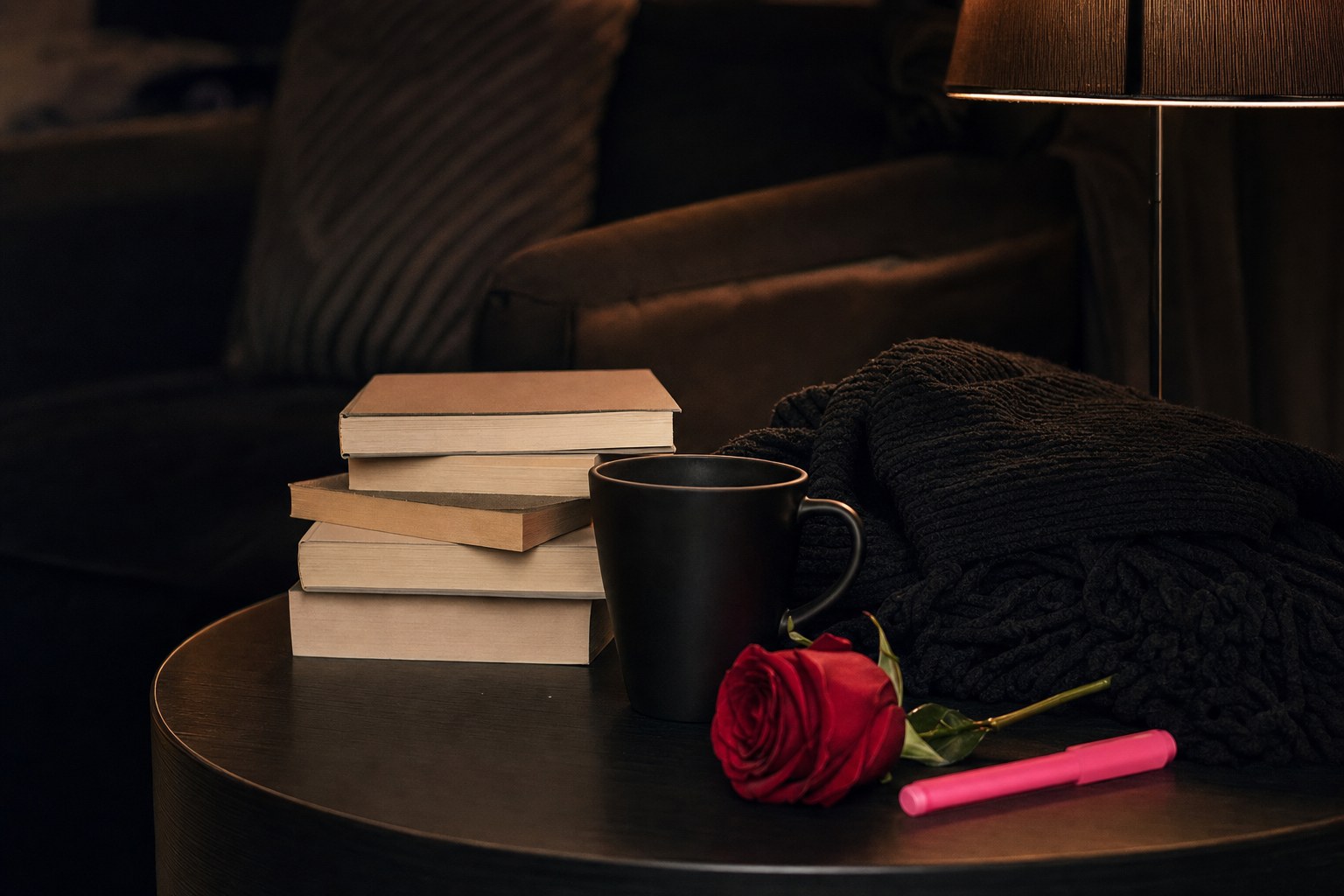 Cozy dark reading nook with a black mug, stacked books, throw, rose, and pink highlighter