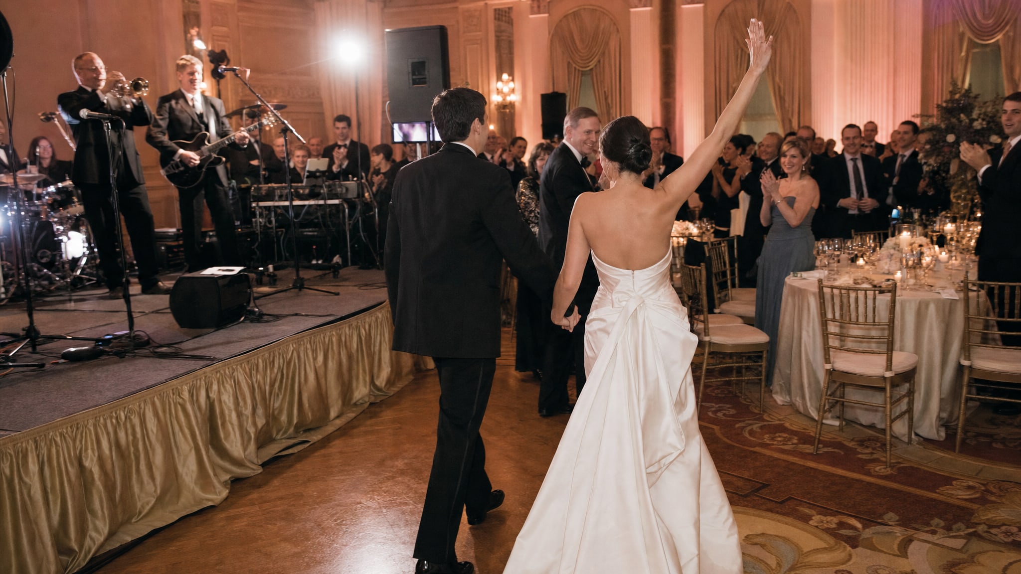 Newlyweds entering a ballroom reception as guests celebrate and a live band performs