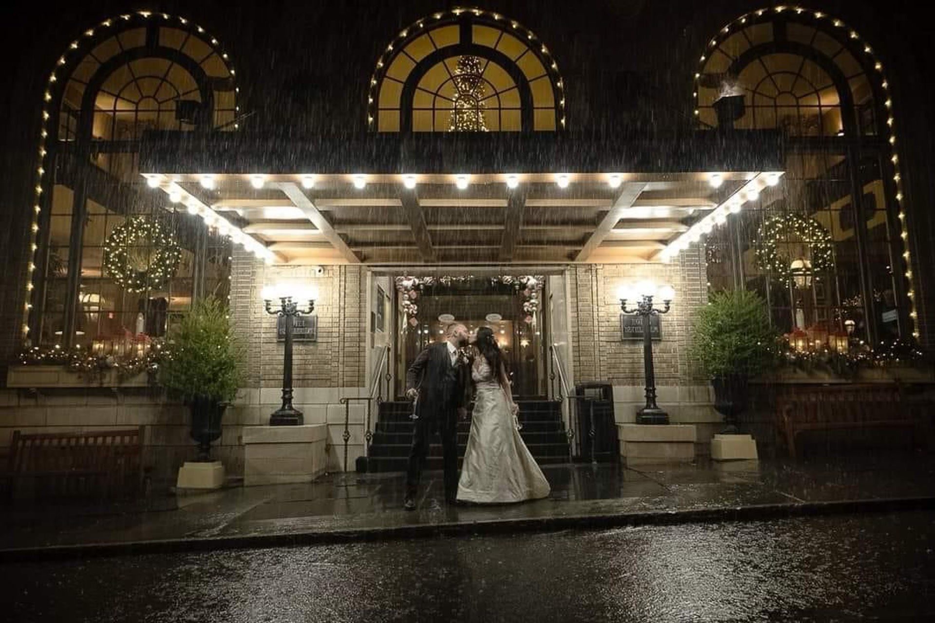 Newlyweds sharing a kiss outside a warmly lit wedding venue in the rain